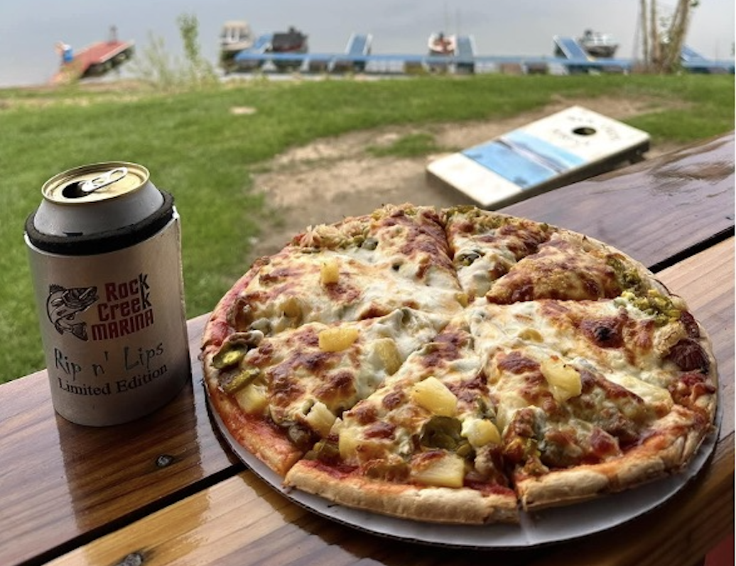 Pizza and Rock Creek Marina branded beer on the lakeside with blue lounge chairs